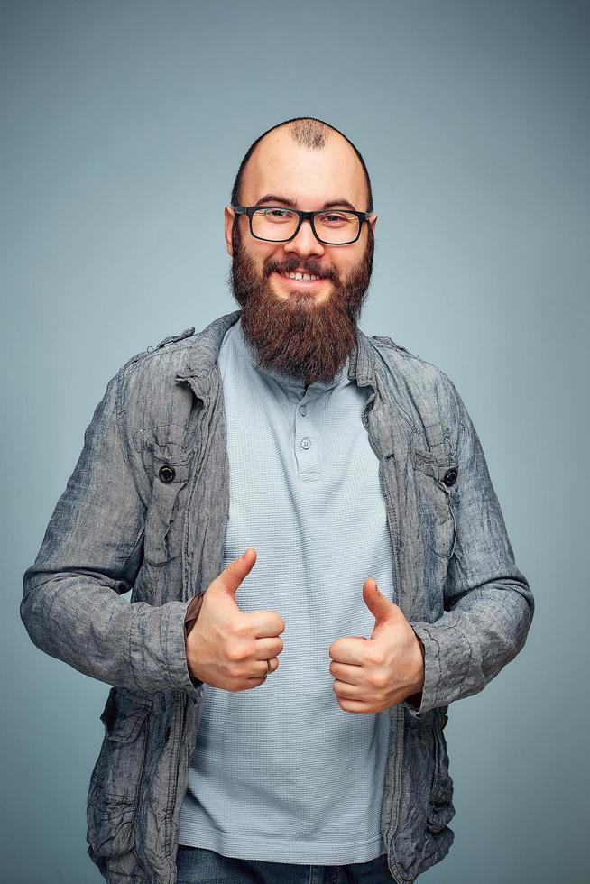 El estilo de vida de un joven exitoso con gafas, barba, chaqueta vaquera de moda mostrando los pulgares hacia arriba, retrato emocional de los hombres en el estudio foto