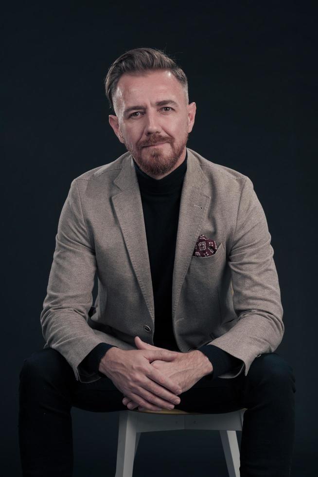 Portrait of adult businessman wearing trendy suit and sitting in modern studio on stylish chair against the black background photo