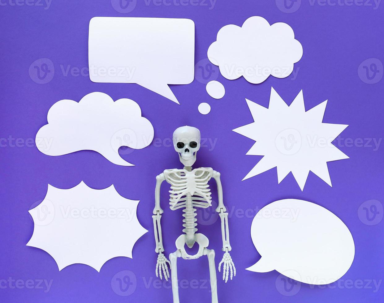 Skeleton on violet background with lots of white blank paper speech bubbles. Anatomical plastic model human skeleton with variety of emotions. Empty dialog clouds, place for text. Purple Halloween. photo