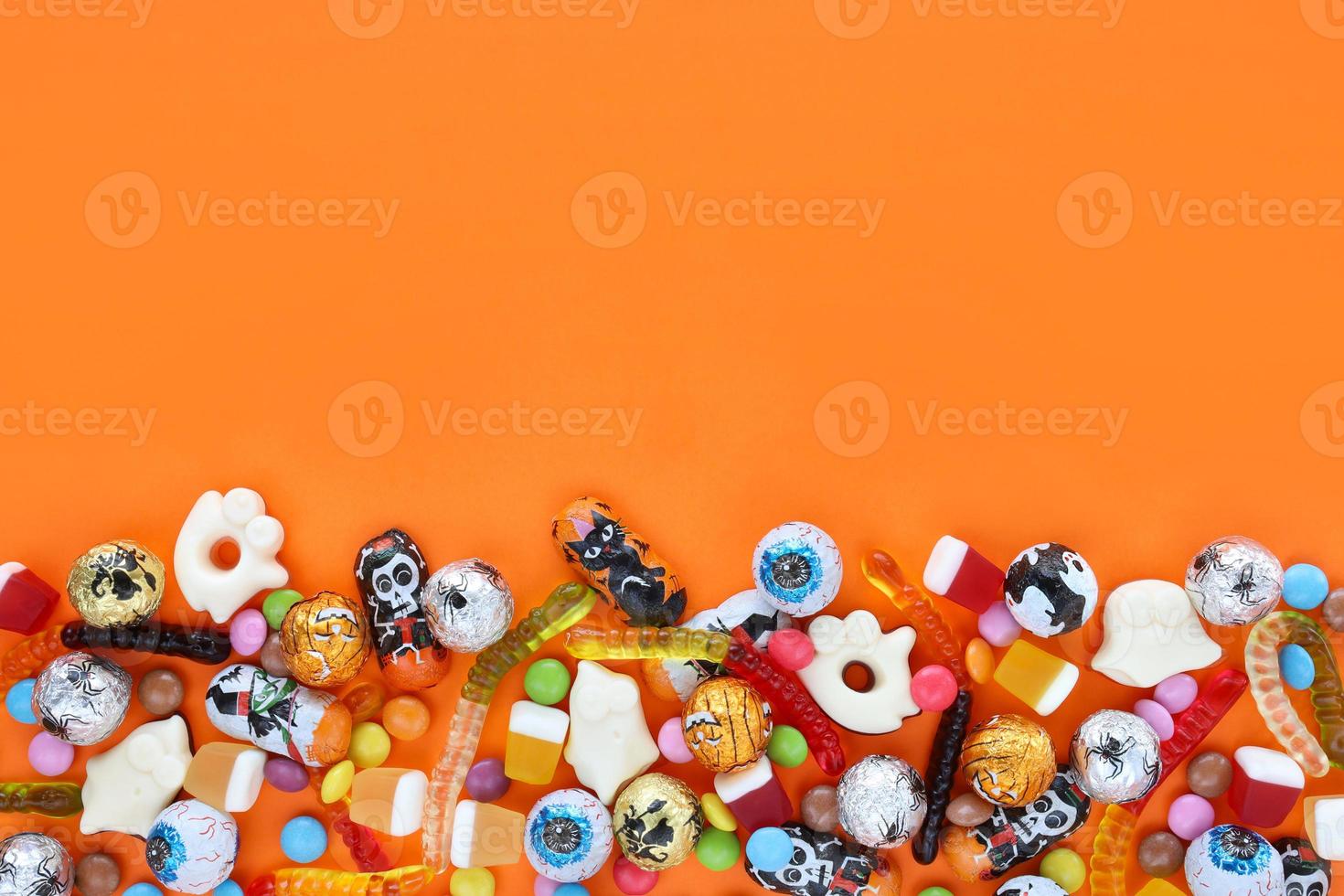 Orange background with copy space, halloween assorted candies at bottom - traditional eyeballs chocolates, jelly worms, ghosts. Happy Halloween holiday sale and trick-or-treat concept. photo