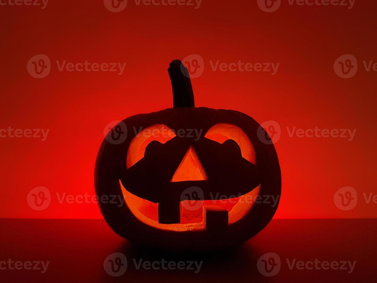 Halloween holiday background with traditional smiling jack-o-lantern. Big carved glowing pumpkin stands on the surface in dark night. The back wall highlighted in red. photo