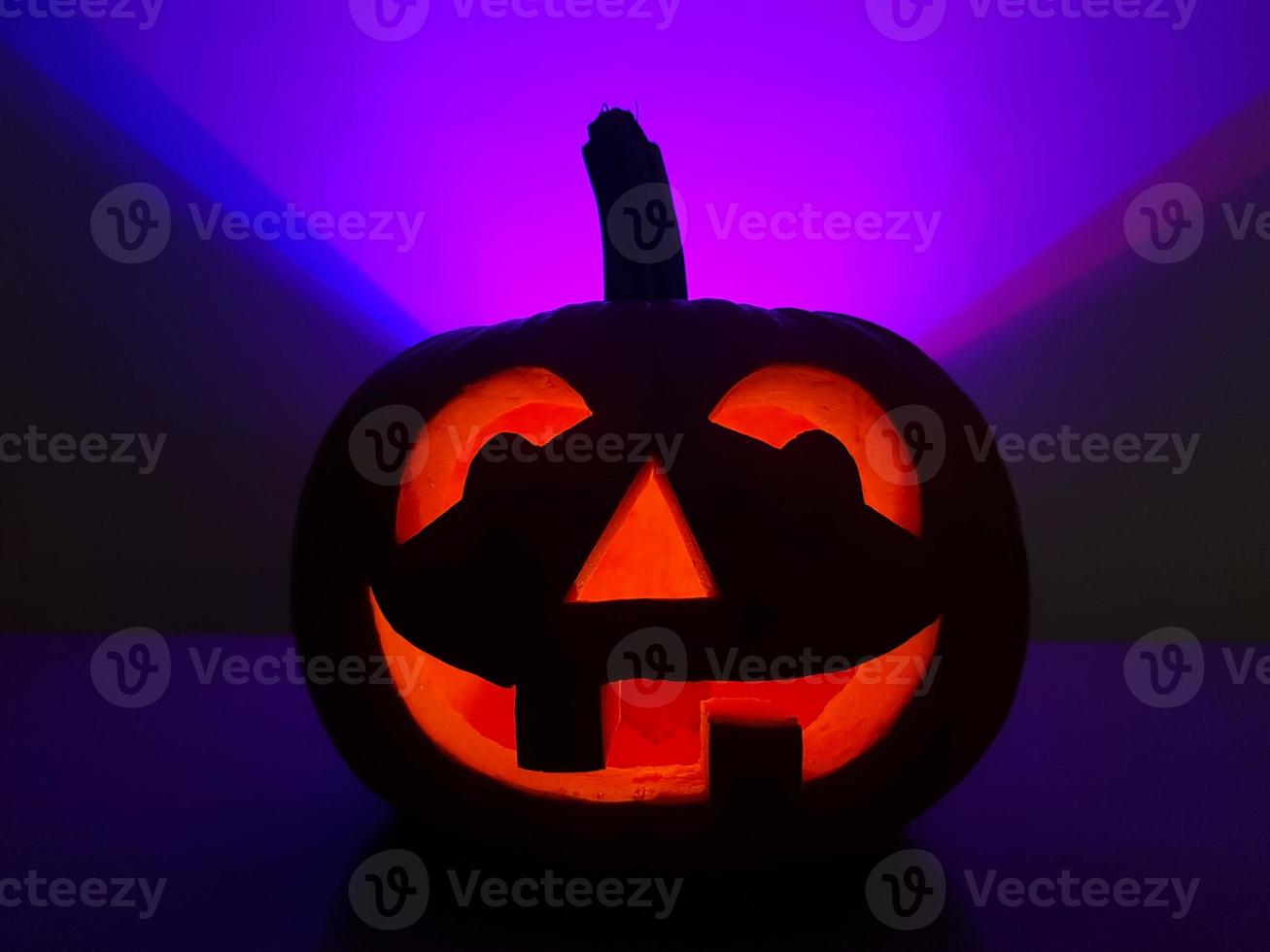 Halloween holiday purple background with orange smiling jack-o-lantern. Big carved glowing pumpkin stands centered on table in dark night. Back wall highlighted in violet triangle over pumpkin. photo