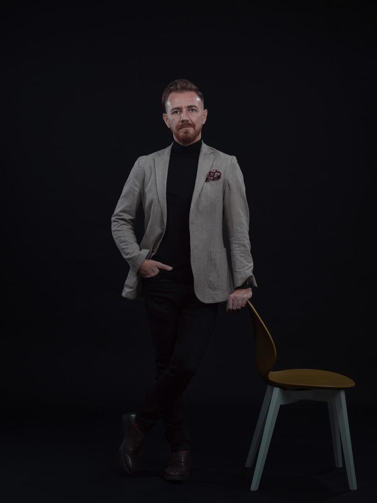 Portrait of adult businessman wearing trendy suit and sitting in modern studio on stylish chair against the black background photo