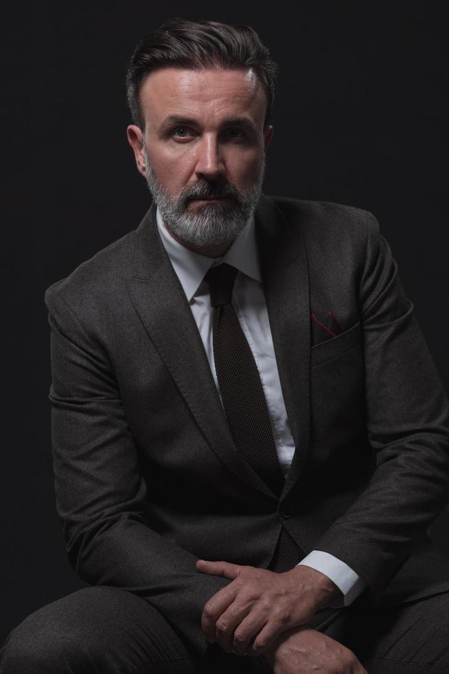 Portrait of adult businessman wearing trendy suit and sitting in modern studio on stylish chair against the black background photo