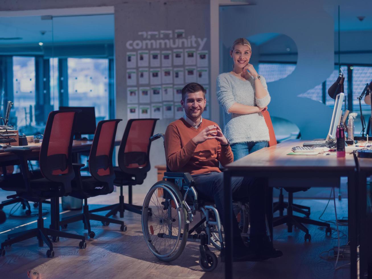 Businessman in a wheelchair in modern coworking office space working late night in office. Colleagues in background. Disability and handicap concept. photo