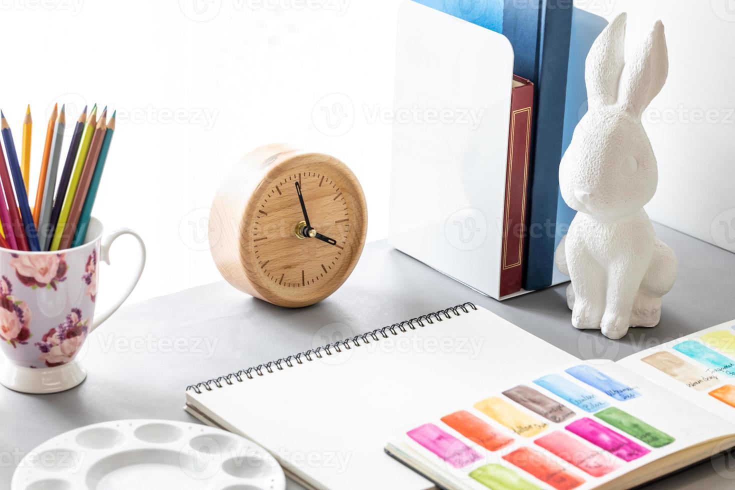 espacio de trabajo del artista de la acuarela con cuaderno de bocetos en blanco, paleta, tubo de color, lápices de colores, reloj de madera y estatua de conejo blanco, vista lateral sobre fondo gris. foto