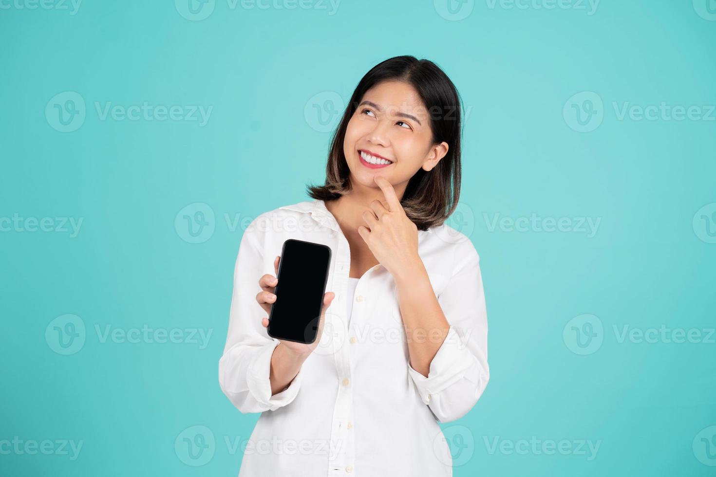 una joven asiática sonriente con ropa informal de pie sostiene un teléfono móvil con una pantalla vacía en blanco que se burla del espacio de copia aislado en un fondo verde foto