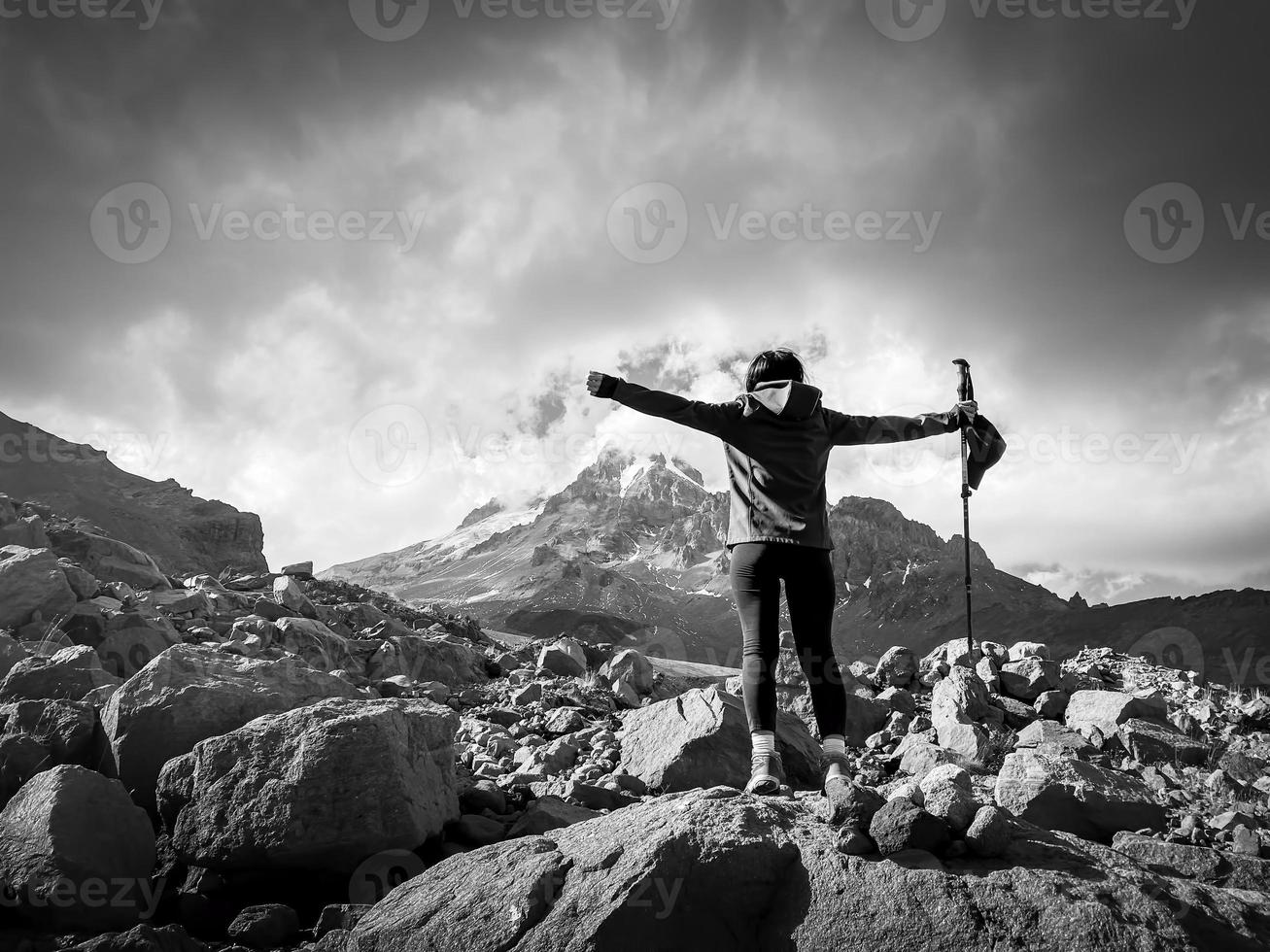 Woman hiker on rock stand on viewpoint with hands up spreaded enjoy panorama of KAzbek mountain in cloudy dramatic day photo