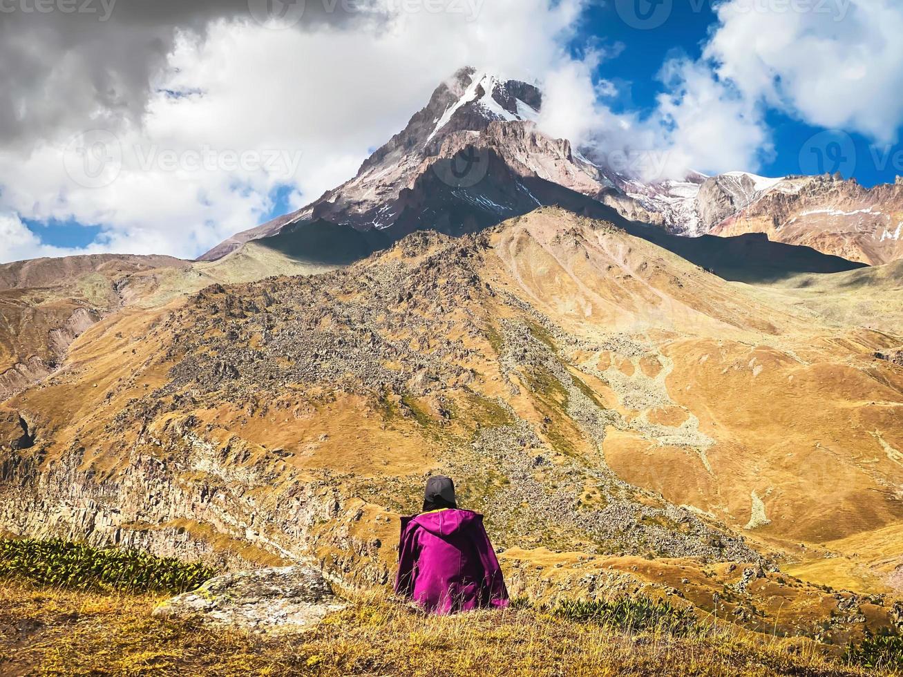 Thoughtful woman sit on grass admire beautiful KAzbek mountain snowy peak in autumn. Hike to gergeti glacier route solo. Travel in caucasus concept photo