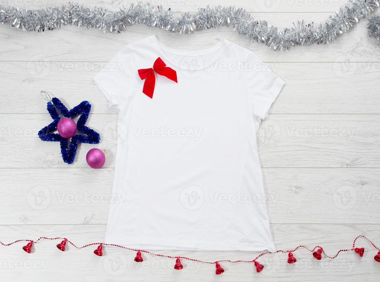 Close up white blank template t shirt with copy space and Christmas Holiday concept. Top view mockup t-shirt and red bow on white wooden background. Happy New Year decorations accessories. Xmas outfit photo
