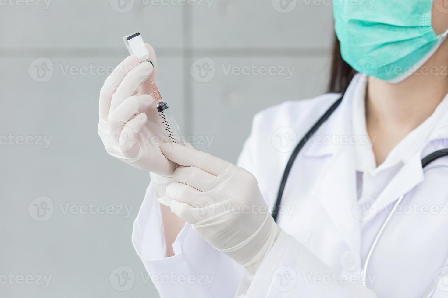 Woman doctor is using syringe to suck vaccine Coronavirus vaccine from bottle while her hands wear rubber gloves and face mask. photo