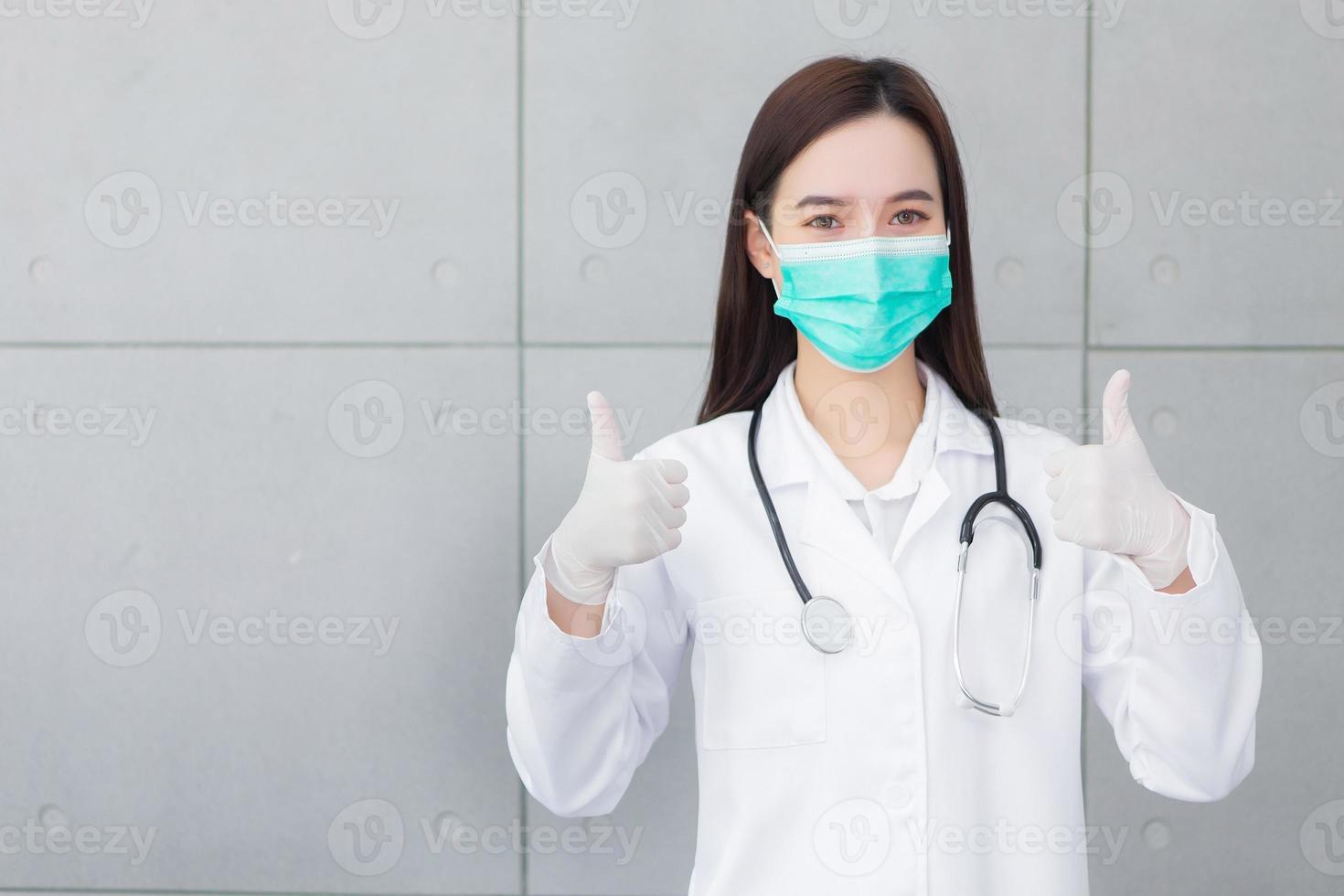 Asian professional woman doctor who wears medical coat and face mask shows thump up as good sign in health protection concept at hospital. photo