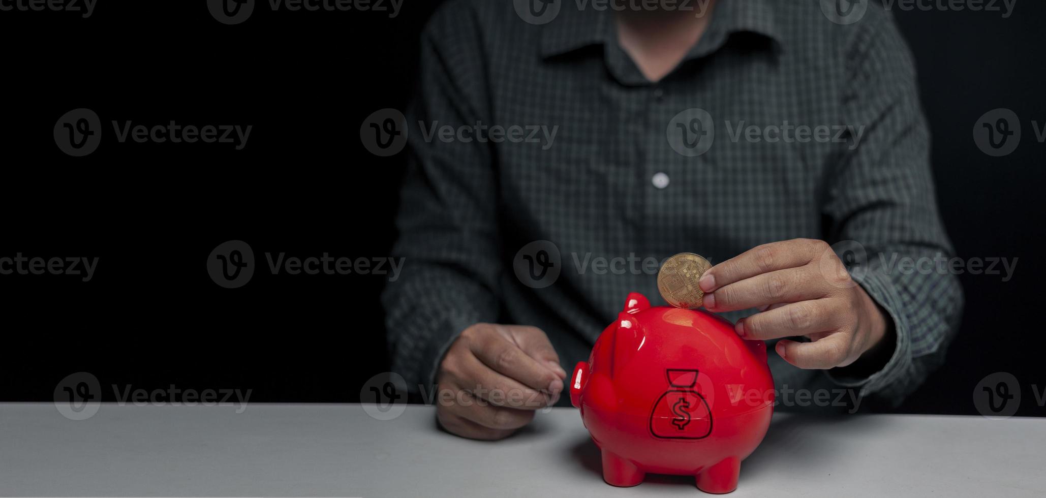 A man's hand is dropping digital gold coins into a red piggy bank on a table inside a dark room with black walls and empty spaces. photo