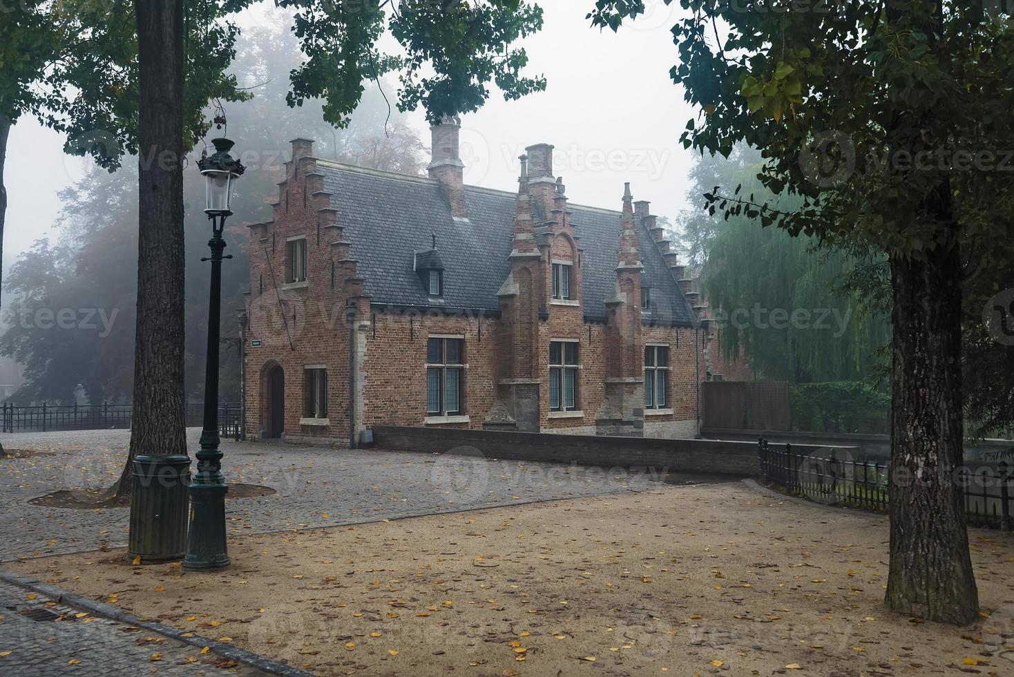 Lonely misty autumn morning on the old medieval city park in Bruges, Belgium. Ancient brick house and cobblestone pavement with fallen leaves wide angle view. photo