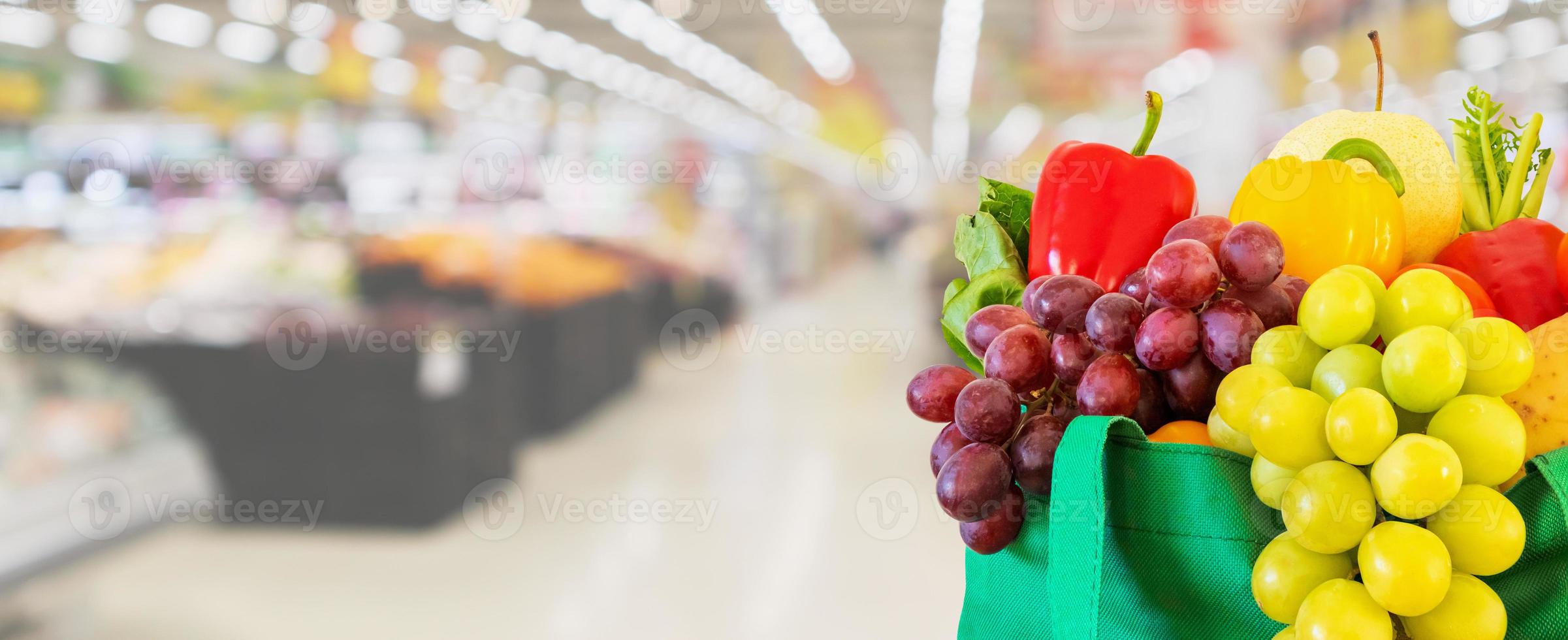 Fresh fruits and vegetables in reusable green shopping bag with supermarket grocery store blurred defocused background with bokeh light photo