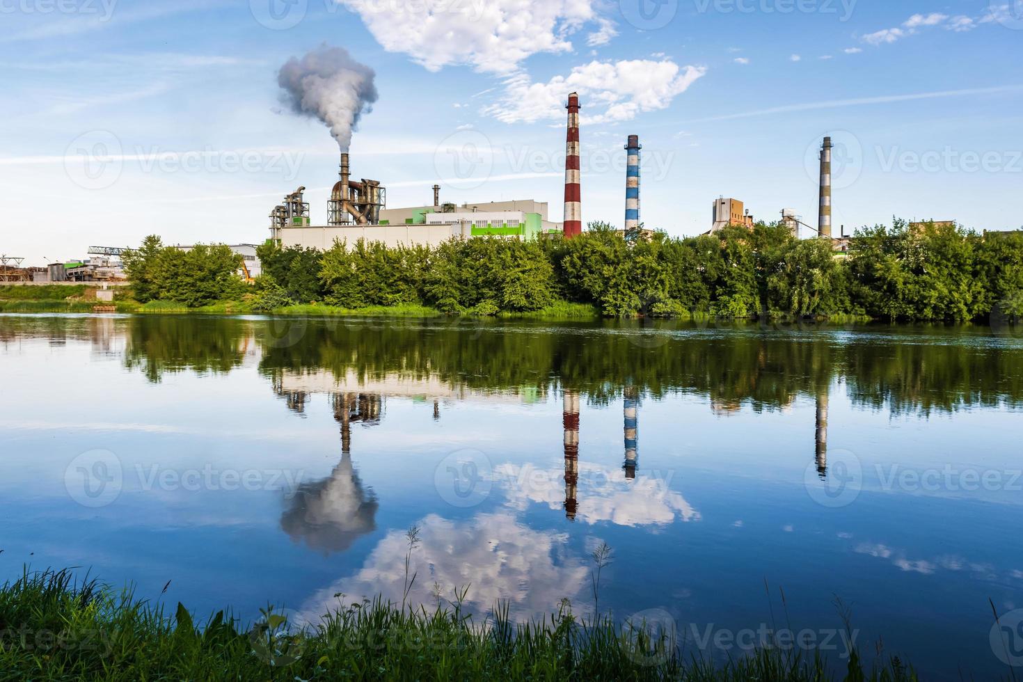 tuberías de aserradero de planta empresarial de carpintería con un hermoso reflejo en el agua azul del río. concepto de contaminación del aire. paisaje industrial contaminación ambiental residuos de central térmica foto