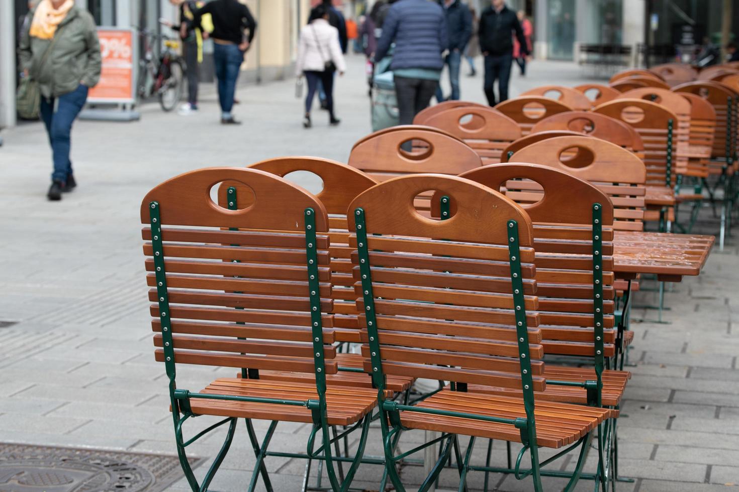 Empty wooden chairs and tables stand in the middle of the pedestrian zone. The furniture is wet from the rain. People in heavy clothing walk past it. photo