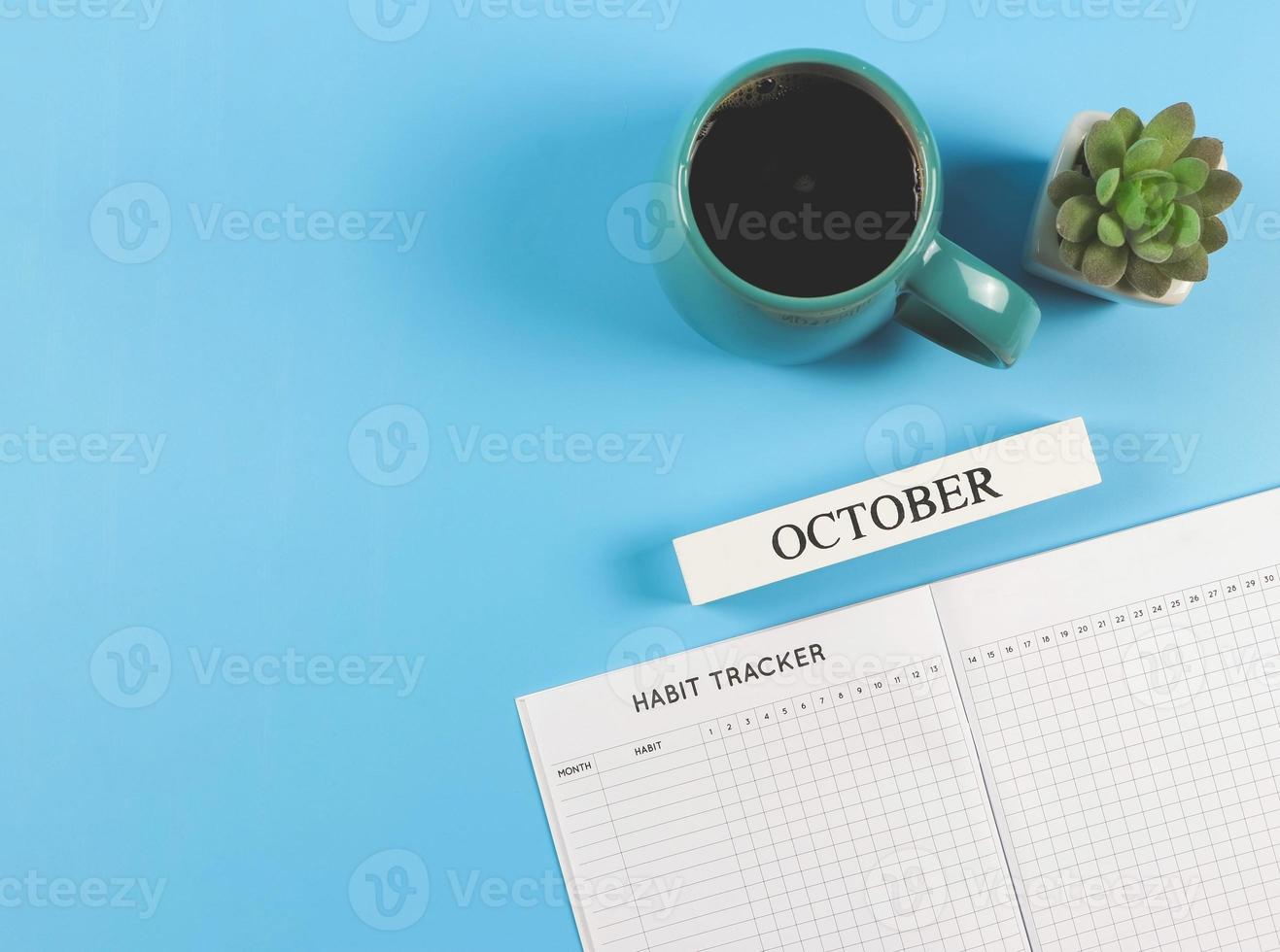 flat lay of habit tracker book, wooden calendar October,  blue cup of black coffee and succulent plant pot on blue background with copy space. photo