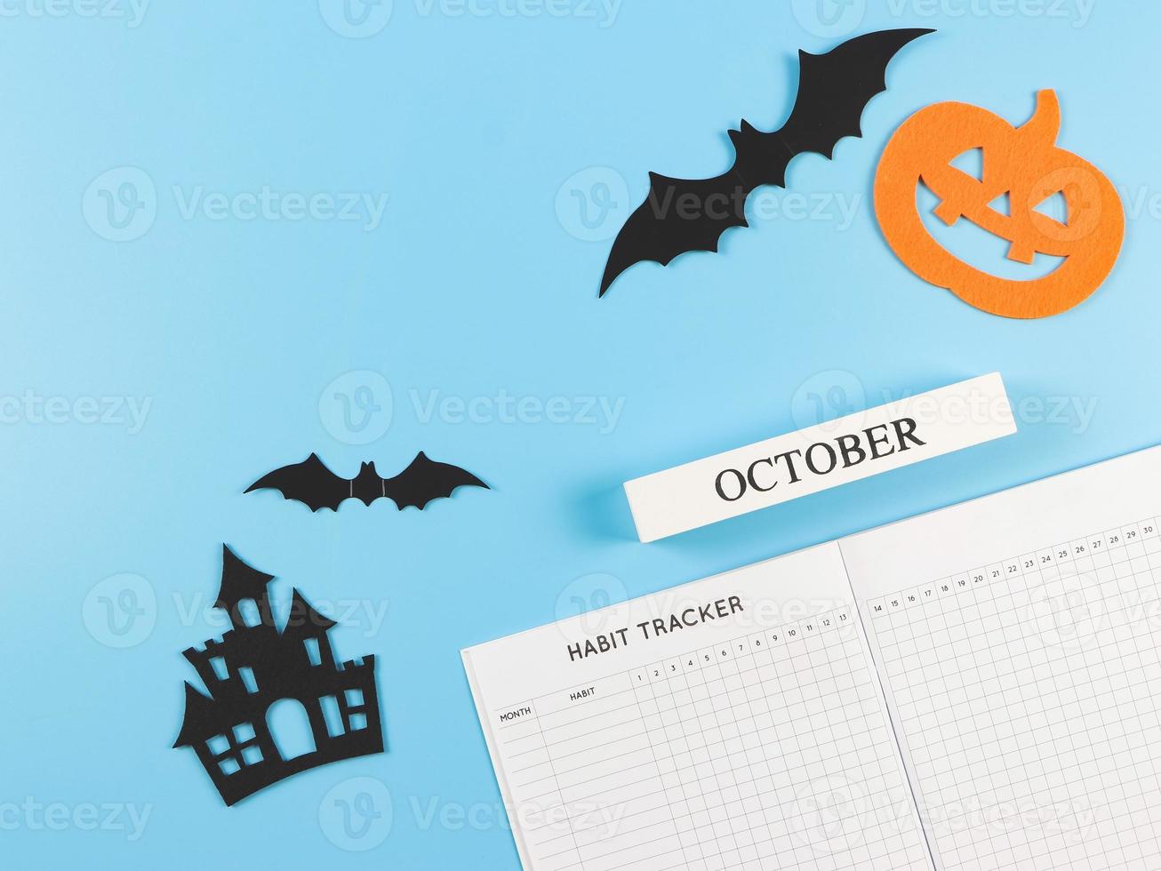 flat lay of habit tracker book, wooden calendar October,  halloween decorations, pumpkin, castles and bats on blue background with copy space. photo