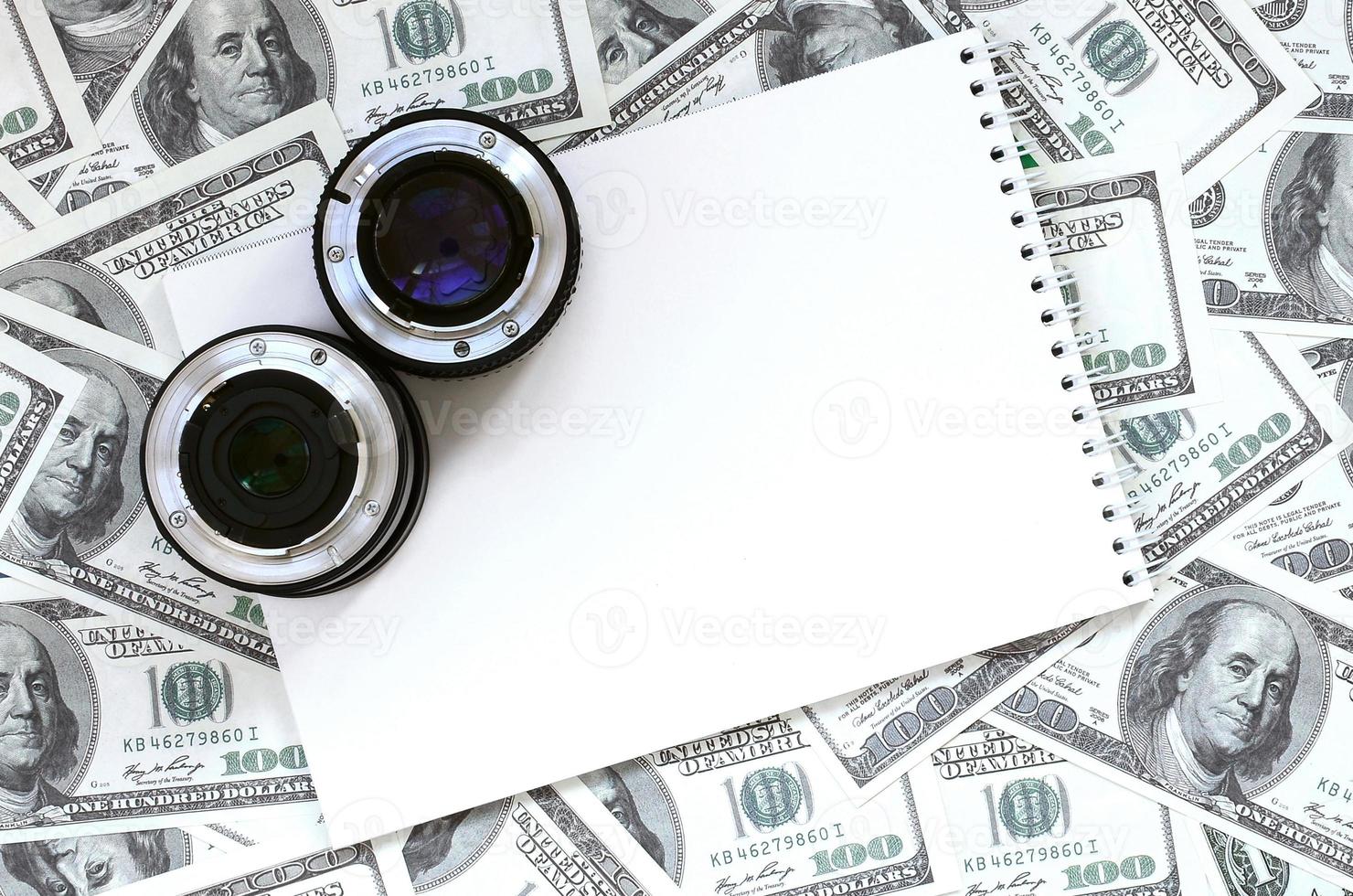 dos lentes fotográficos y un cuaderno blanco se encuentran en el fondo de muchos billetes de dólar. espacio para texto foto