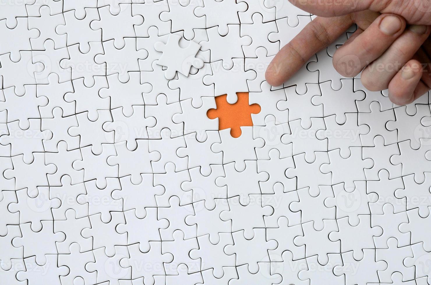 The texture of a white jigsaw puzzle in the assembled state with one missing element, forming an orange space, pointed to by the finger of the male hand photo