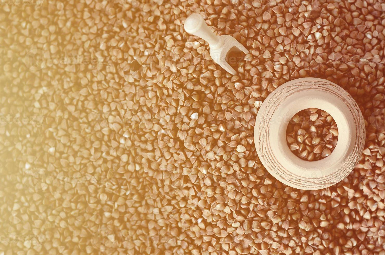 Background image of a large pile of buckwheat, in the middle of which lies a small jug and a wooden spatula for cereals photo