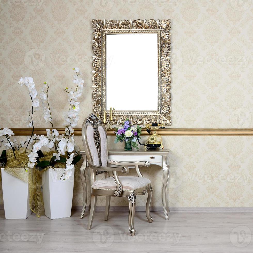 A fragment of a retro interior with a chair and table, on which is a telephone and a vase of flowers. Above the table hangs an empty picture frame for your photo