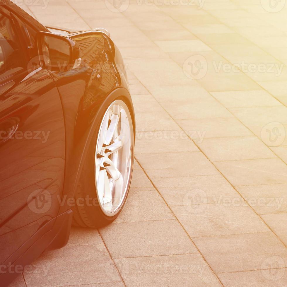 Diagonal view of a black glossy car with white wheels, which stands on a square of gray tiles photo