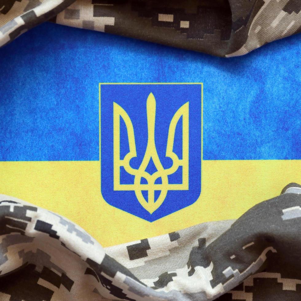 SUMY, UKRAINE - MARCH 20, 2022 Ukrainian flag and coat of arms with fabric with texture of pixeled camouflage. Cloth with camo pattern in grey, brown and green pixel shapes with Ukrainian trident sign photo