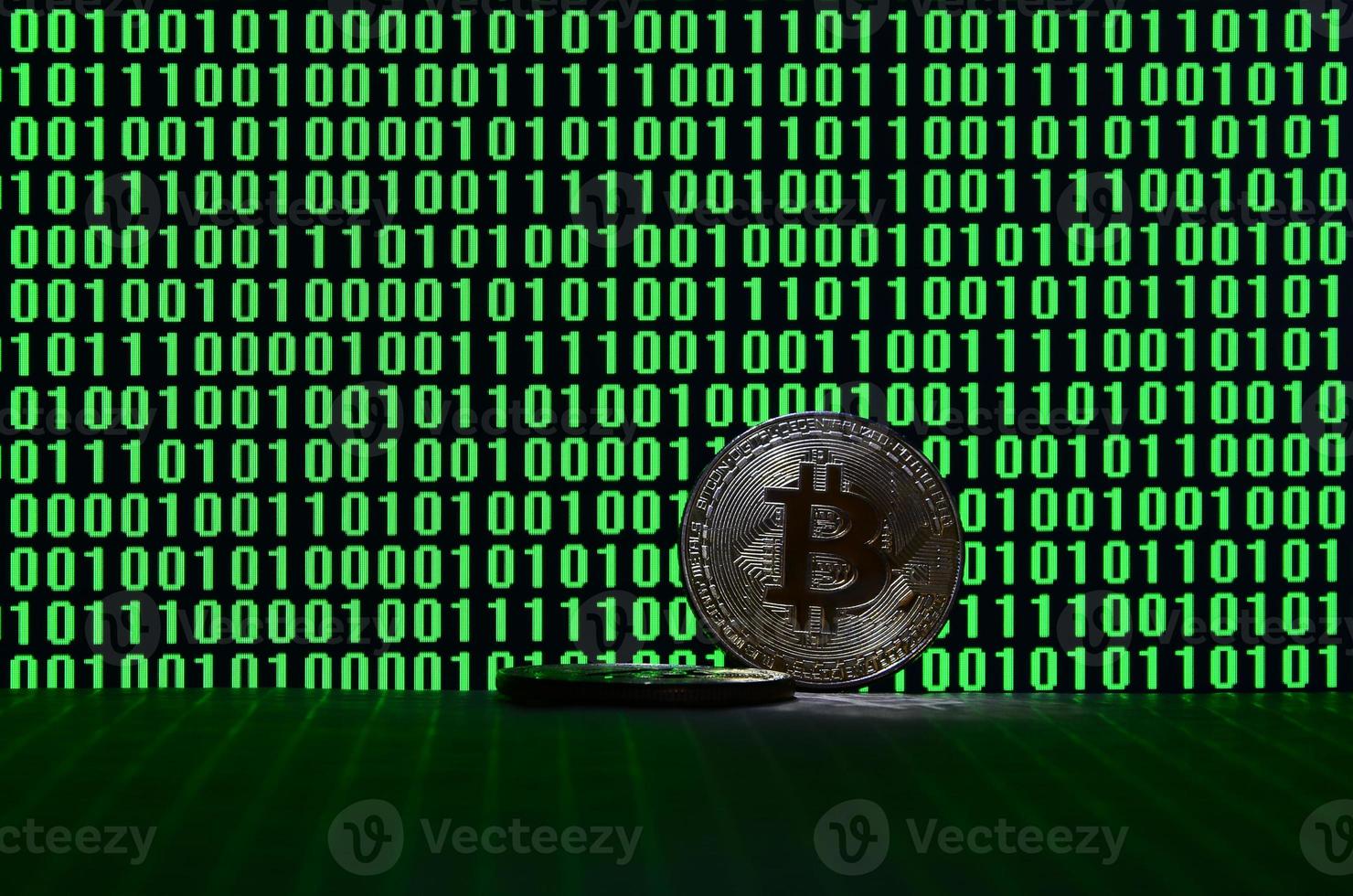 A pair of bitcoins lies on a cardboard surface on the background of a monitor depicting a binary code of bright green zeros and one units on a black background. Low key lighting photo