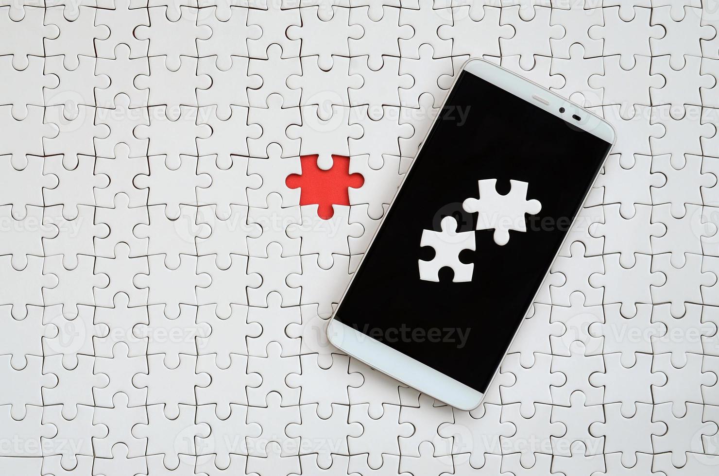 A modern big smartphone with several puzzle elements on the touch screen lies on a white jigsaw puzzle in an assembled state with missing elements photo