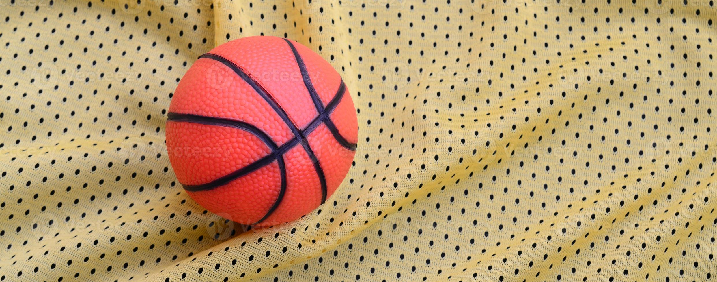 Small orange rubber basketball lies on a yellow sport jersey clothing fabric texture and background with many folds photo