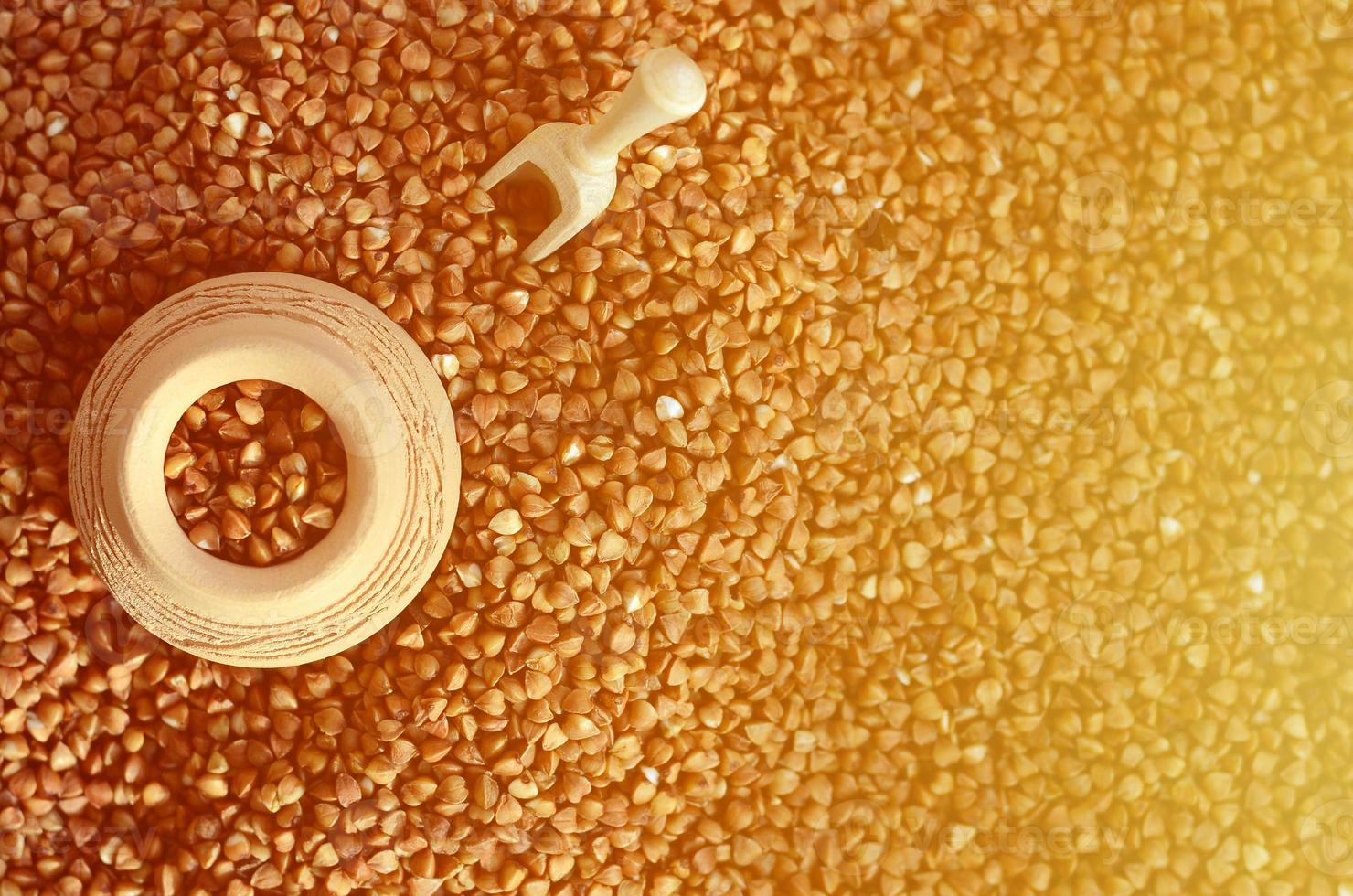 Background image of a large pile of buckwheat, in the middle of which lies a small jug and a wooden spatula for cereals photo