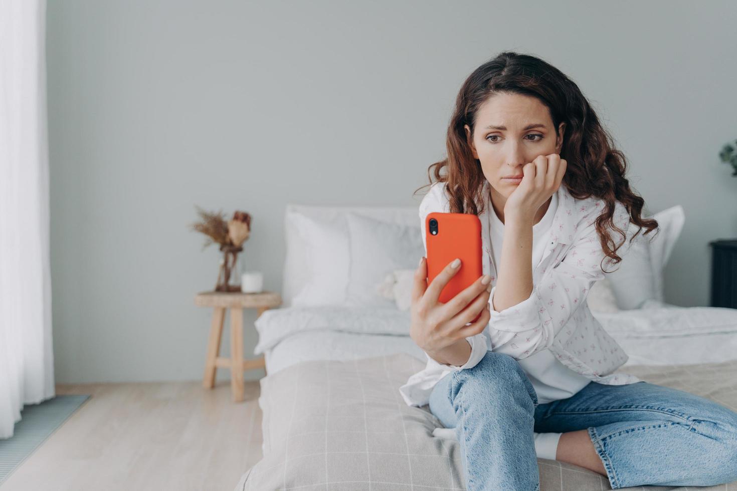 Upset puzzled female reading bad media news in social networks holding smartphone sitting in bedroom photo