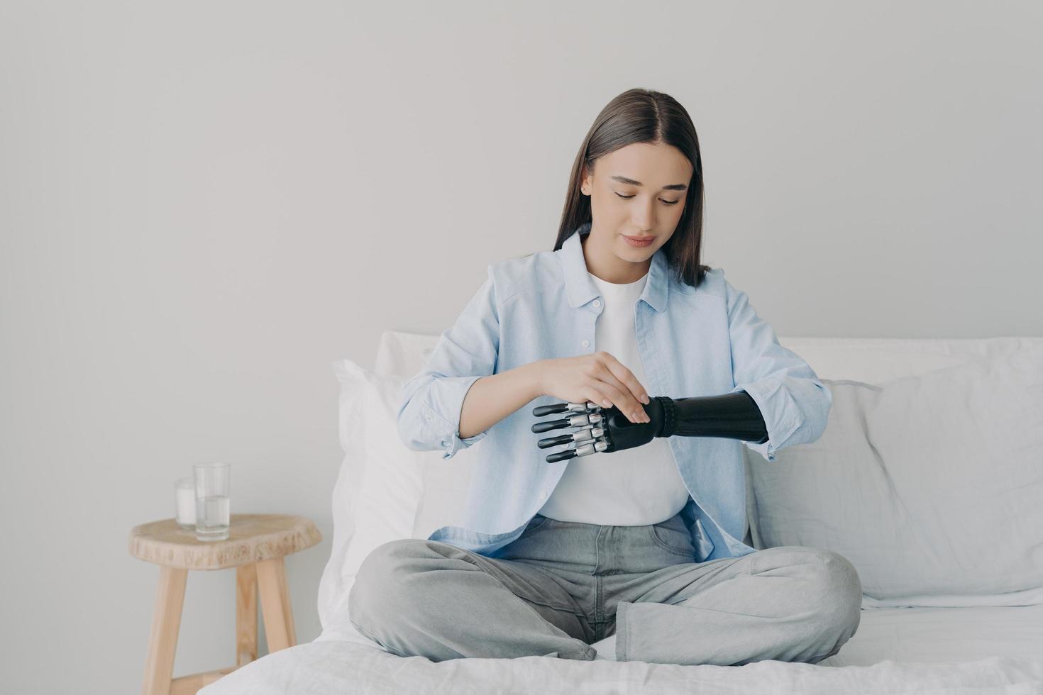 Disabled girl setting bionic arm prosthesis, sitting on bed. Lifestyle of people with disabilities photo