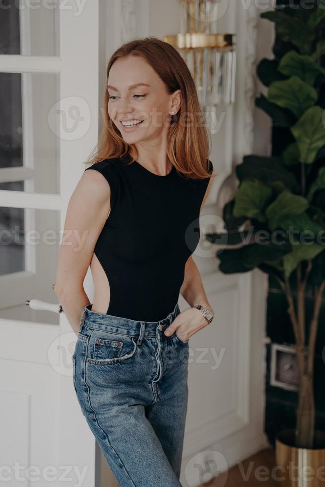 Attractive european lady in fashionable slim fit black bodysuit and jeans looking aside and smiling. photo