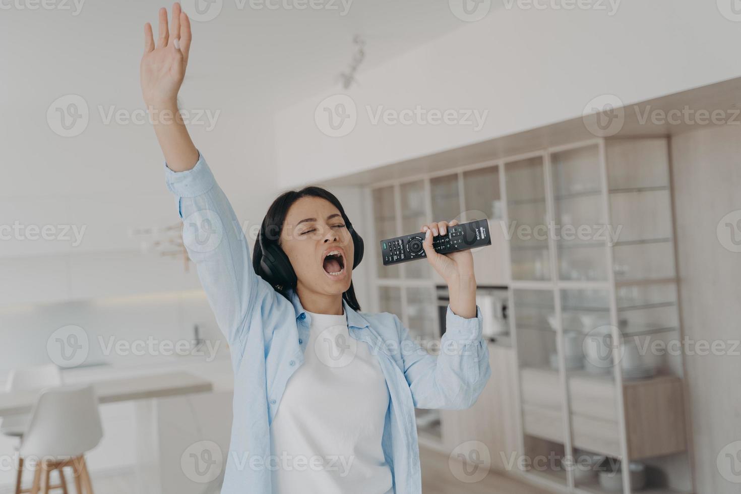 Energetic female in headphones sing karaoke, using TV remote control like a mic, rests at home photo