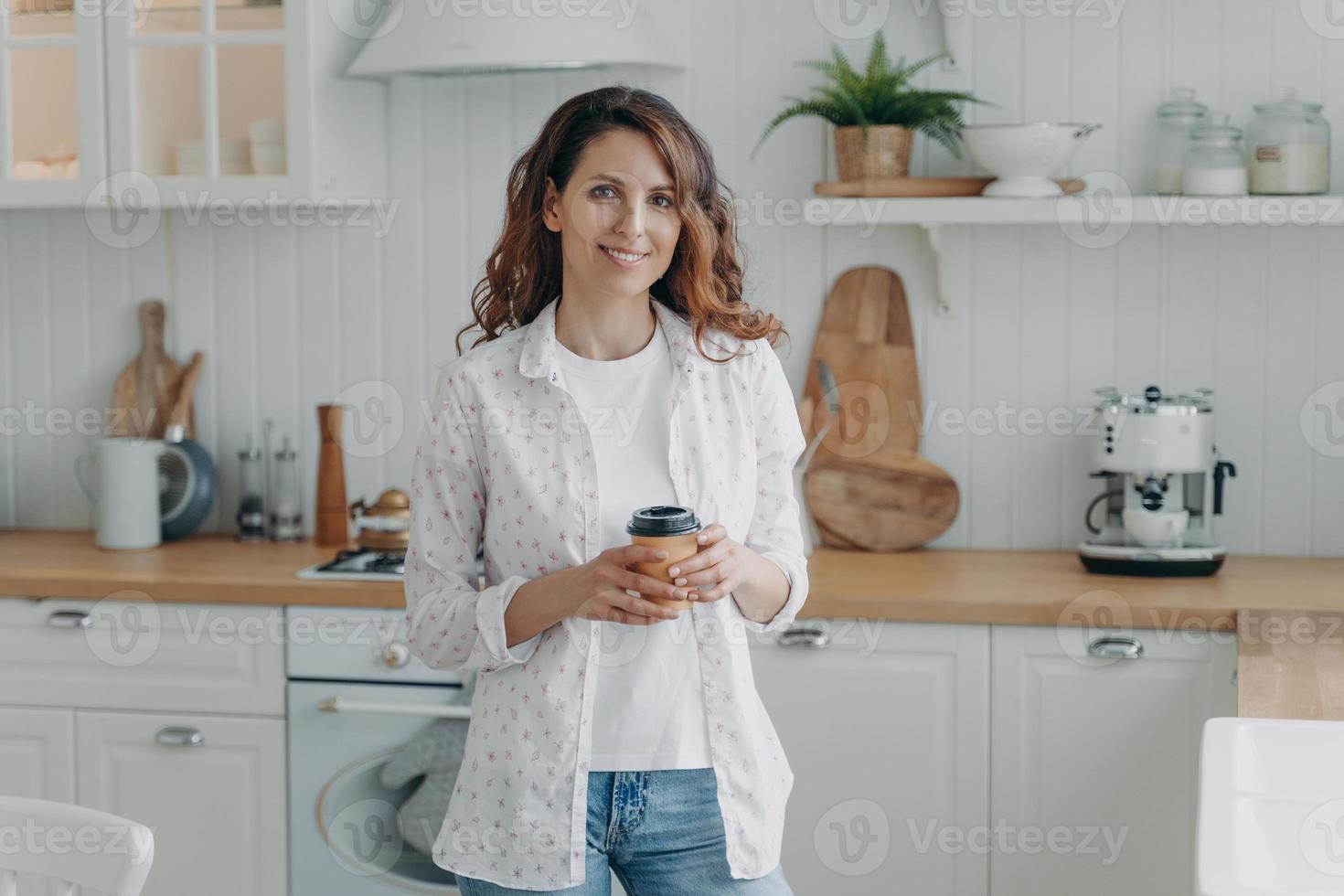 Smiling girl hold paper cup of coffee, standing in modern cozy kitchen look at camera. Domestic life photo