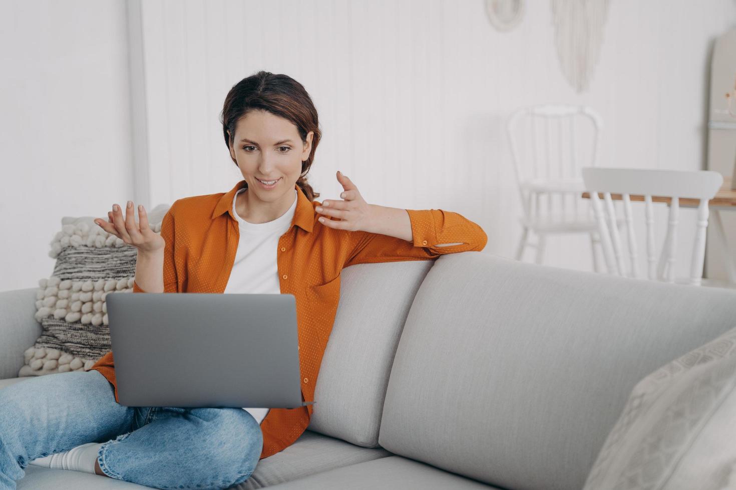 chica sorprendida charlando por videollamada en la computadora portátil, asombrada con buenas noticias, sentada en el sofá en casa foto