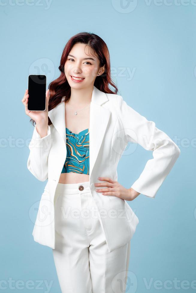 Half body of confident elegant beautiful Asian Business woman wearing white suit smile and holding mobile phone on isolated blue background. Young shopaholic female on copy space. photo
