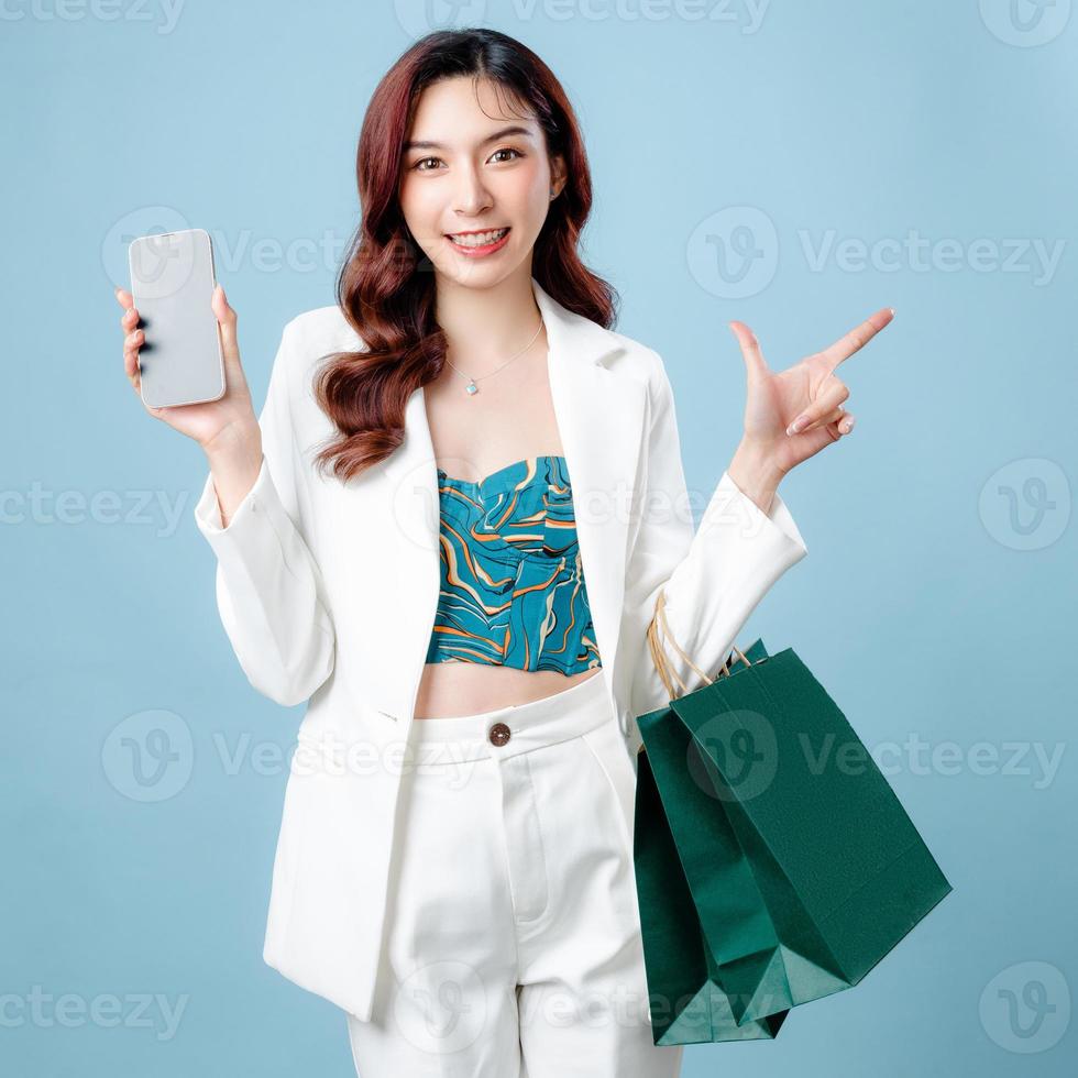 Half body of beautiful Asian Business woman wearing white suit pointing to empty space holding shopping bag and mobile phone on isolated blue background. Blank screen smartphone concept. photo