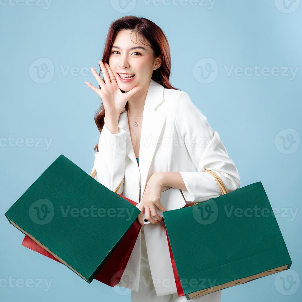 Half body of beautiful Asian Business woman wearing white suit holding shopping bag and mobile phone on isolated blue background. Blank screen smartphone concept. photo