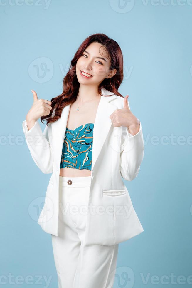 Half body of confident elegant beautiful Asian Business woman wearing white suit smiling and showing thumbs up on isolated blue background. Young shopaholic female on copy space. photo