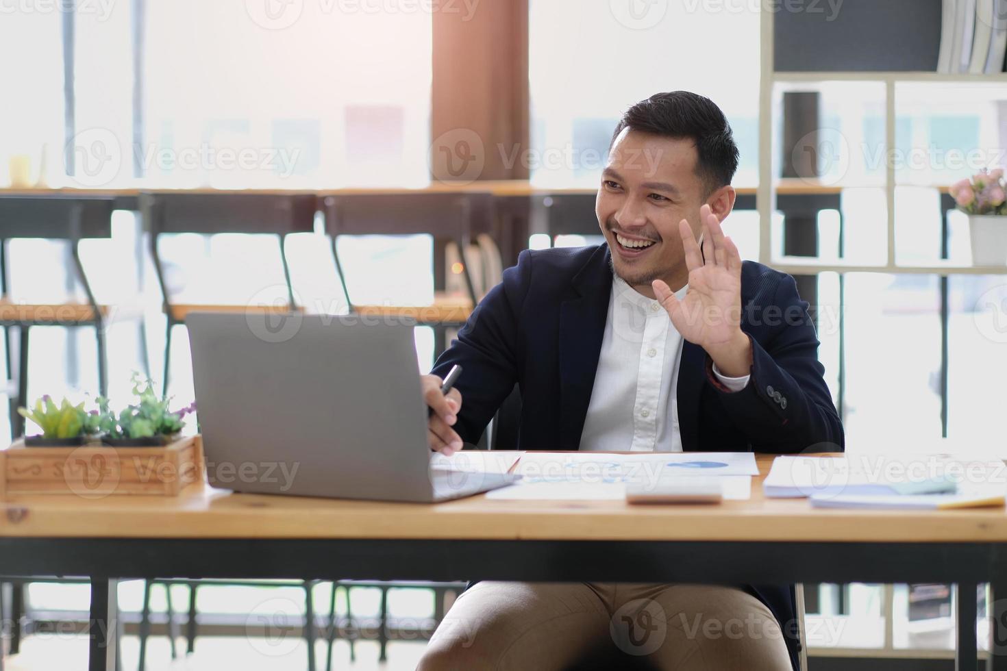 Portrait of a young Asian businessman smiling while using a laptop and writing down notes while sitting at his desk in a modern office photo