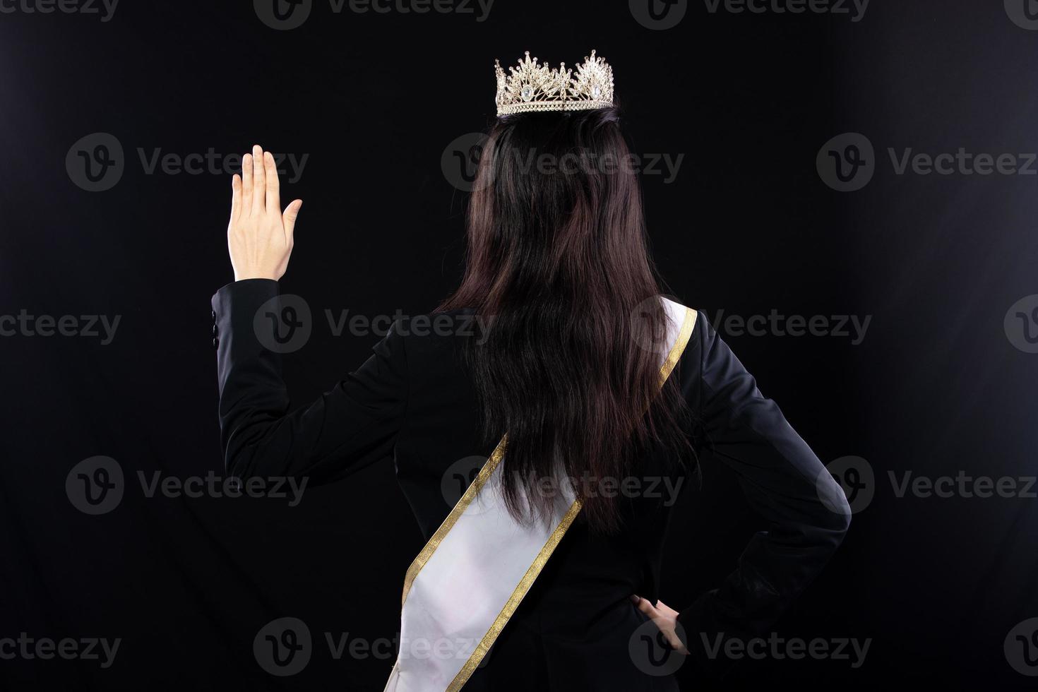 Half body portrait of Miss Pageant Beauty Contest in Asian smart suit blazer shirt dress with Silver Diamond Crown Sash, fashion make up smile wave hand, studio lighting black background isolated photo