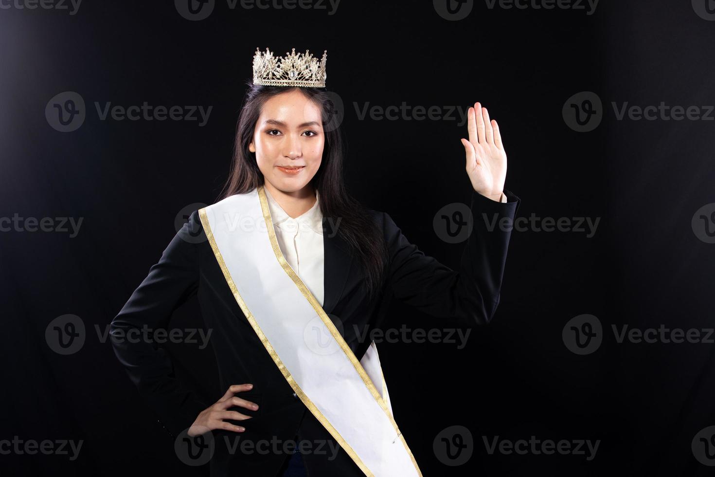 Half body portrait of Miss Pageant Beauty Contest in Asian smart suit blazer shirt dress with Silver Diamond Crown Sash, fashion make up smile wave hand, studio lighting black background isolated photo