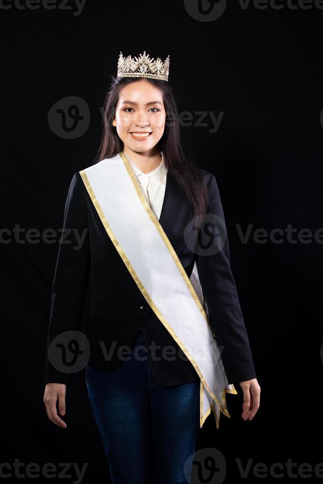 Half body portrait of Miss Pageant Beauty Contest in Asian smart suit blazer shirt dress with Silver Diamond Crown Sash, fashion make up smile wave hand, studio lighting black background isolated photo