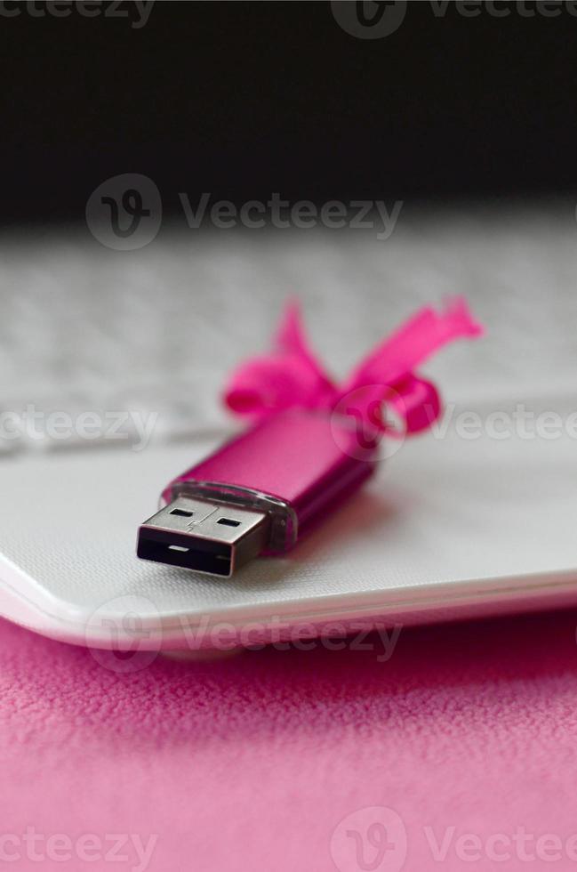 Brilliant pink usb flash memory card with a pink bow lies on a blanket of soft and furry light pink fleece fabric beside to a white laptop. Classic female gift design for a memory card photo