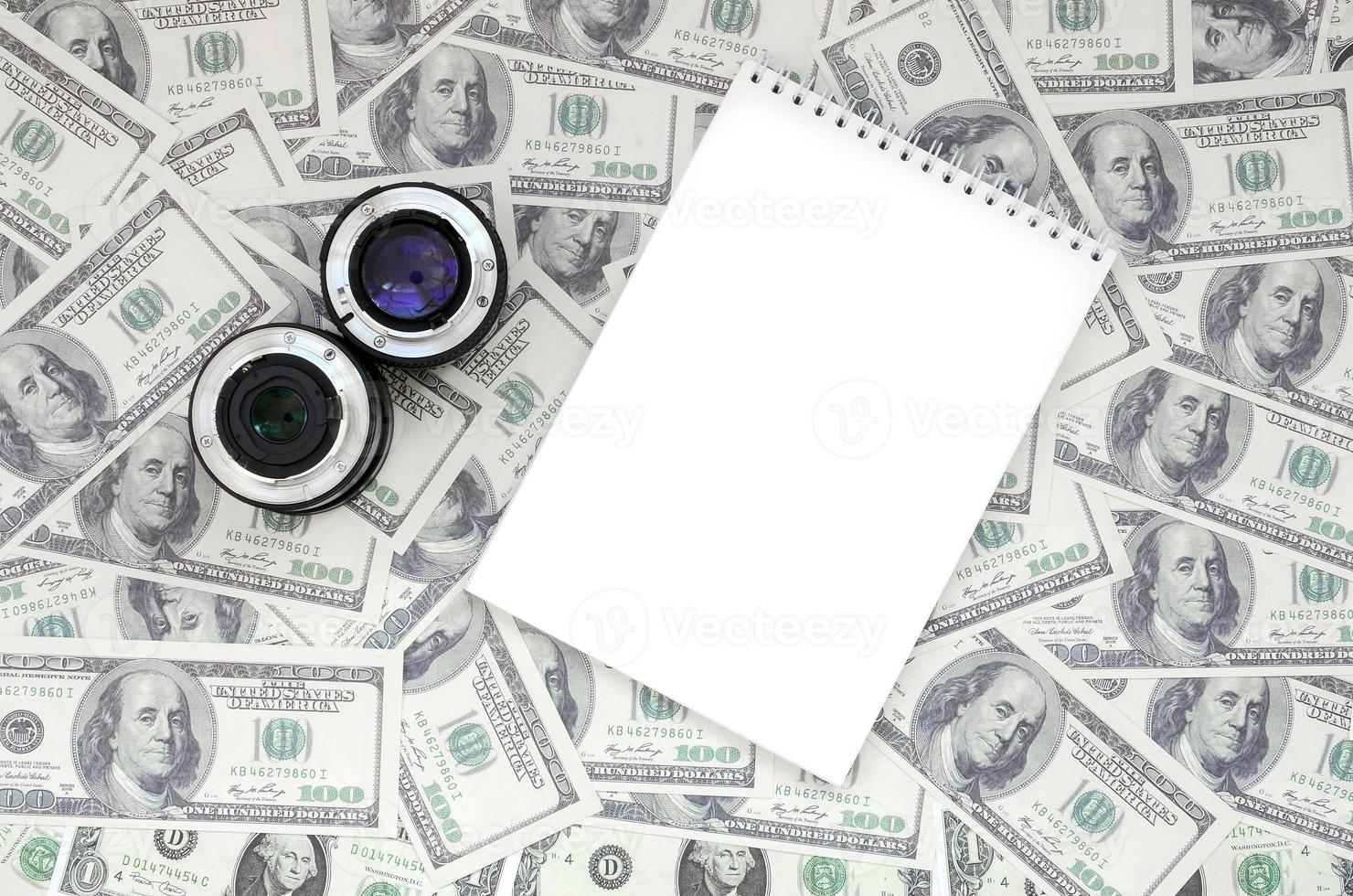 Two photographic lenses and white notebook lie on the background of a lot of dollar bills. Space for text photo
