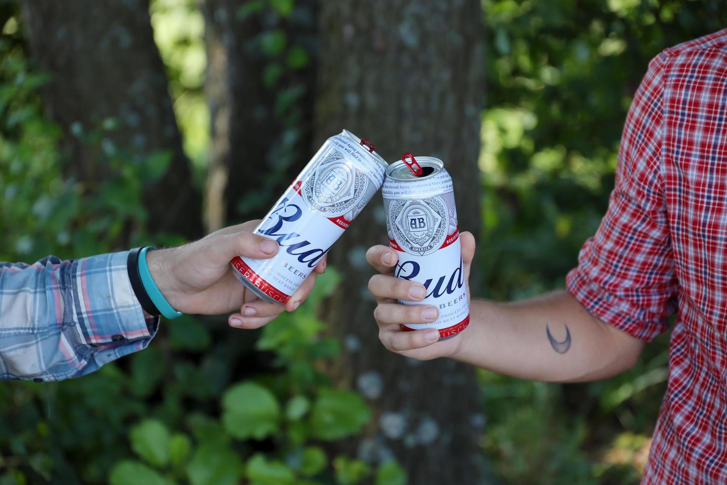SUMY, UKRAINE - AUGUST 01, 2021 Young man raise Budweiser Bud beer can with male friend on blurred river with kayak and trees. Budweiser is one of the most popular beer brands in the USA photo